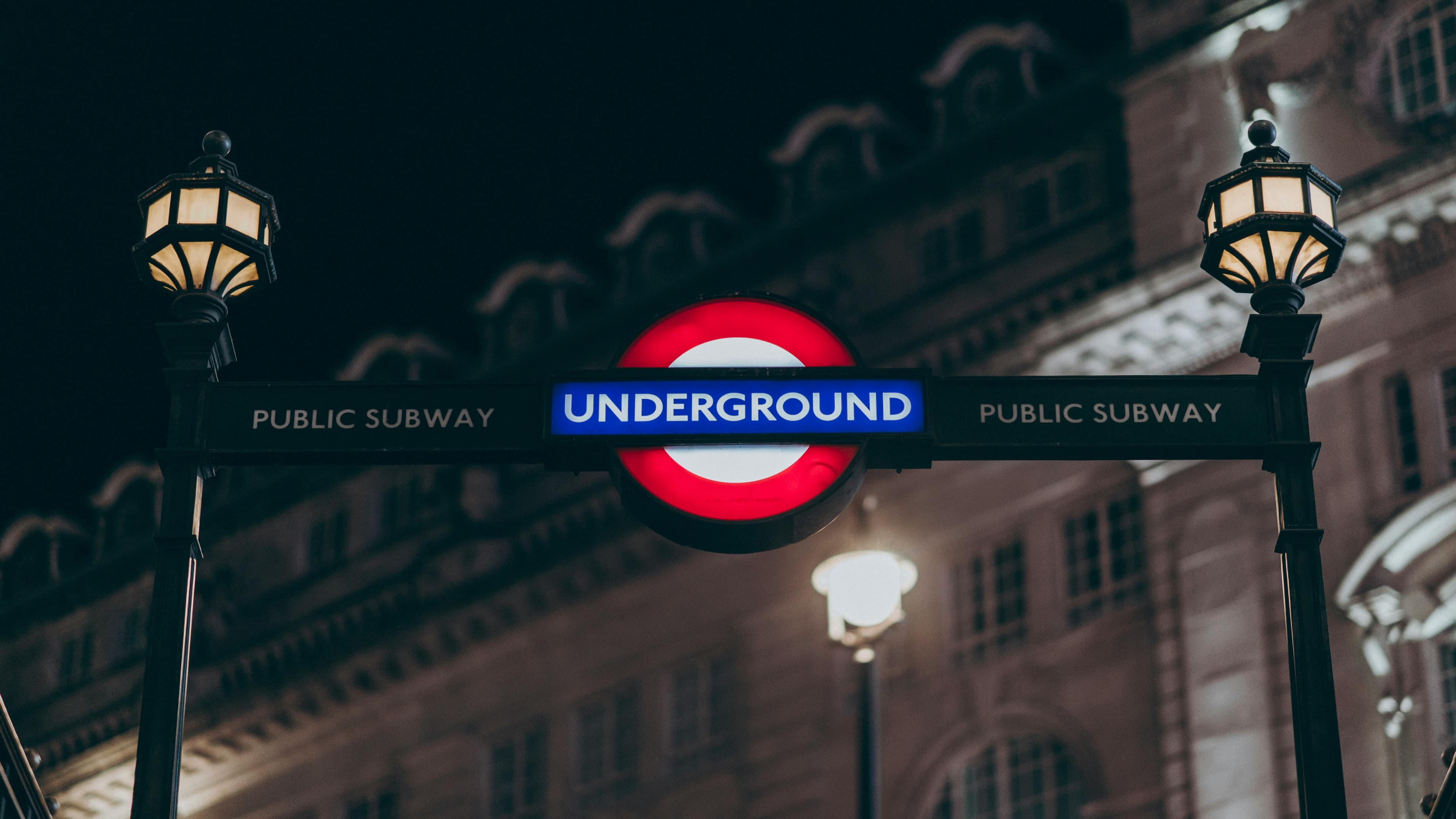 Illuminated London Underground sign flanked by two ornate street lamps against a dark building.