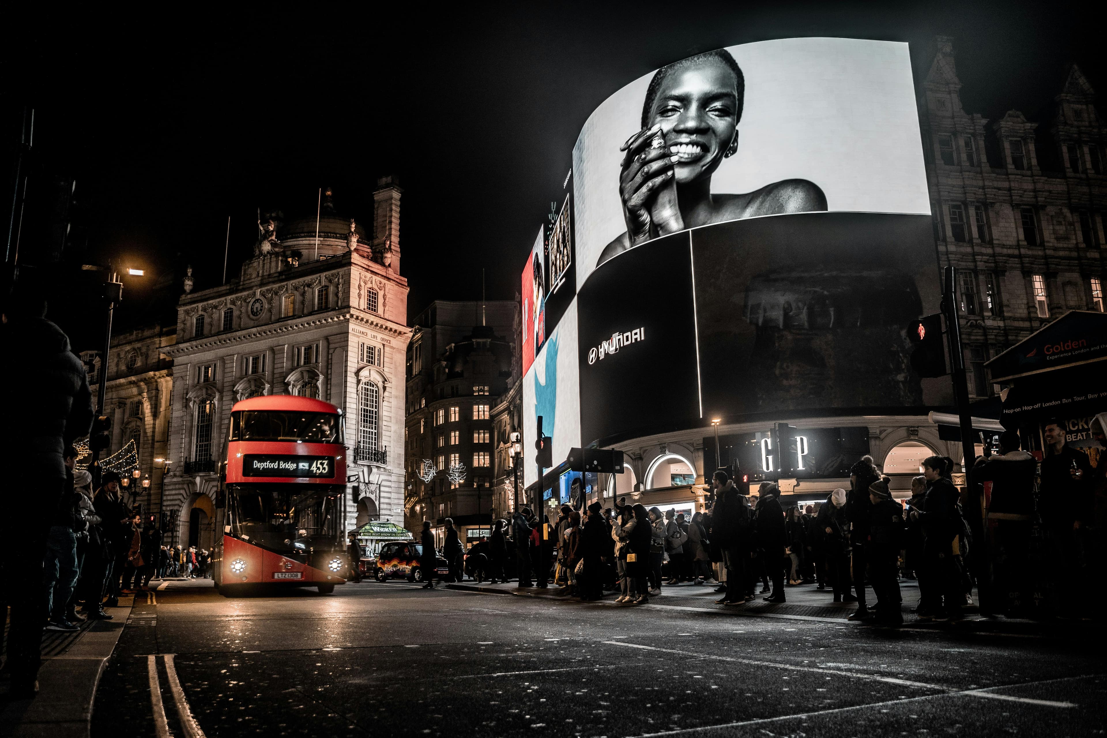 Red double-decker bus passing a massive digital billboard in Piccadilly Circus, London, at night.